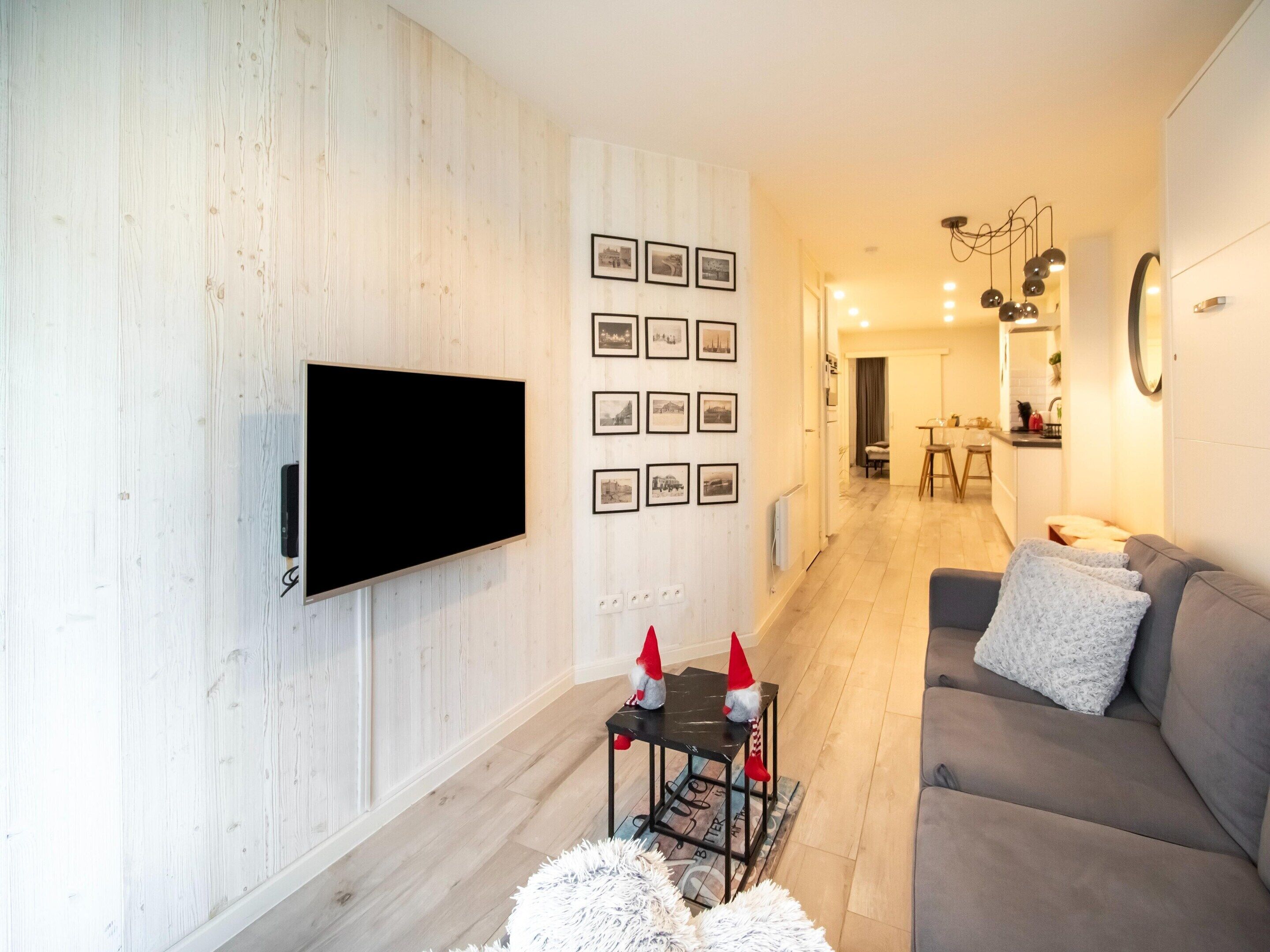 Ein modernes Wohnzimmer mit einem Flachbildfernseher an einer Holzwand, einem grauen Sofa mit einem weißen Zierkissen und einem kleinen Tisch mit dekorativen Gartenzwergen.