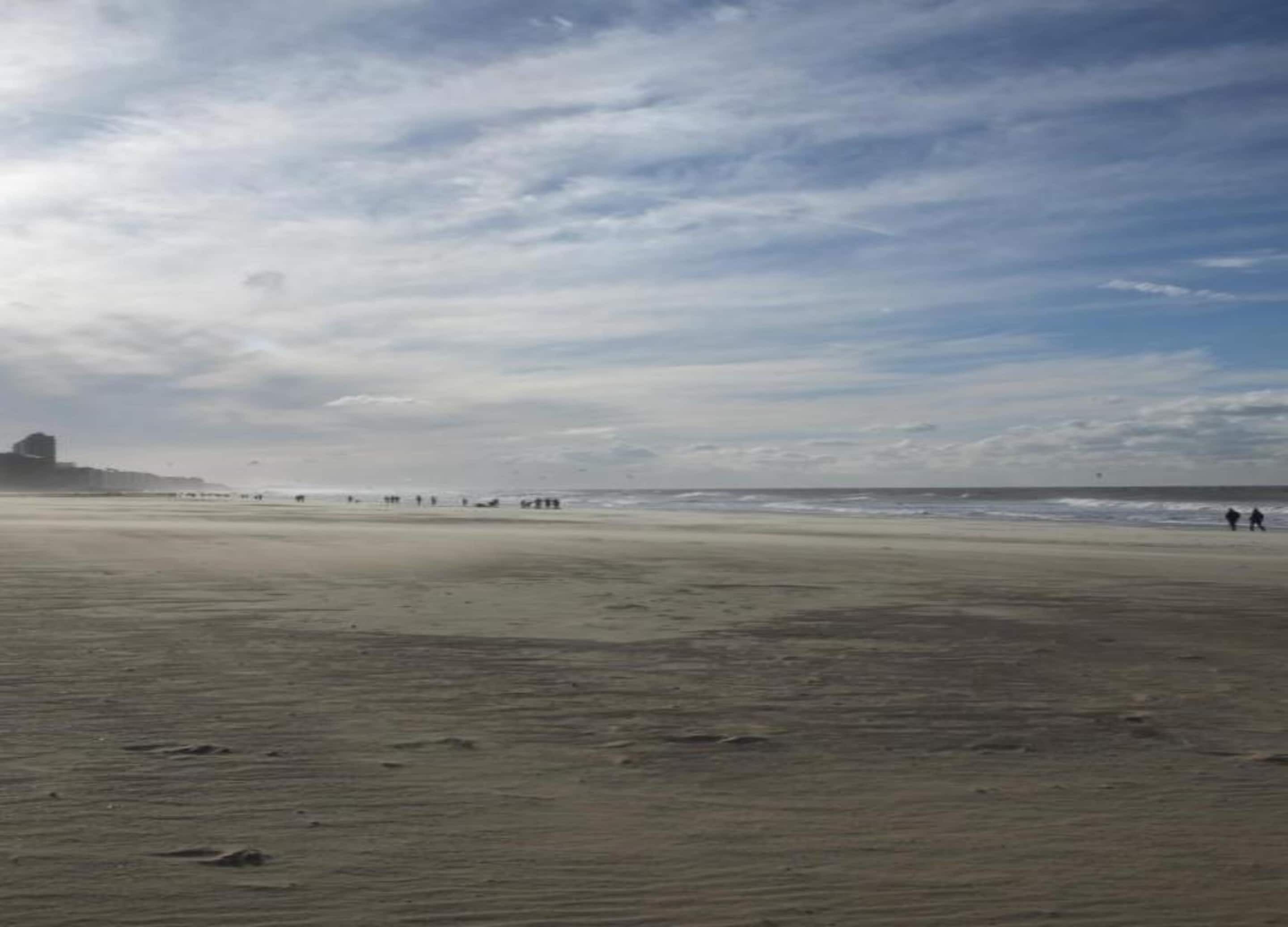 Een breed zandstrand met enkele mensen die wandelen, een gebouw in de verte en een bewolkte lucht.