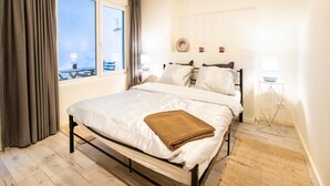 Un dormitorio con cama, ventana, vista al exterior, una mesita de noche con lámpara y estante con objetos decorativos.