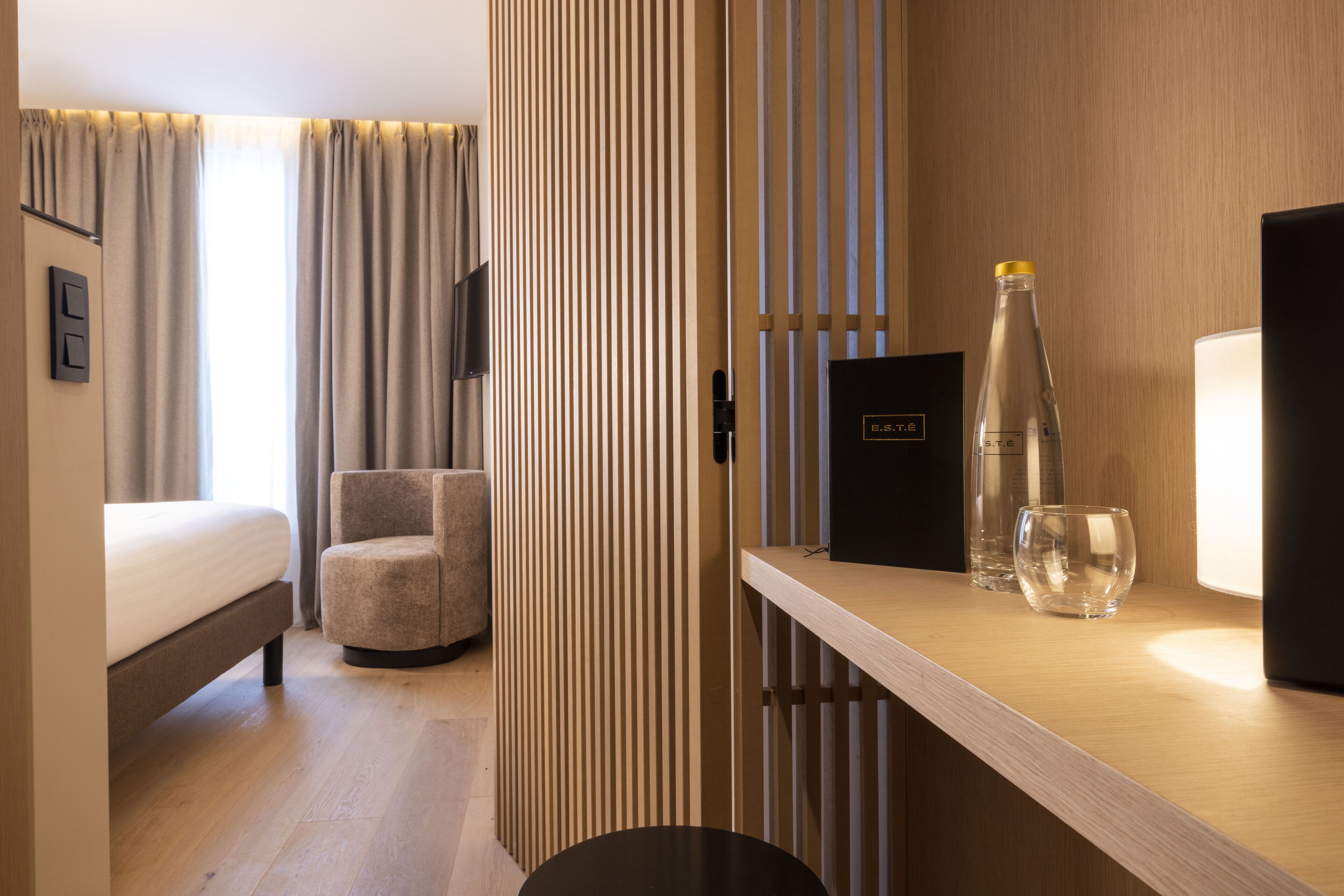 A hotel room with a bed, a chair, a desk with a bottle and glass, and a TV.