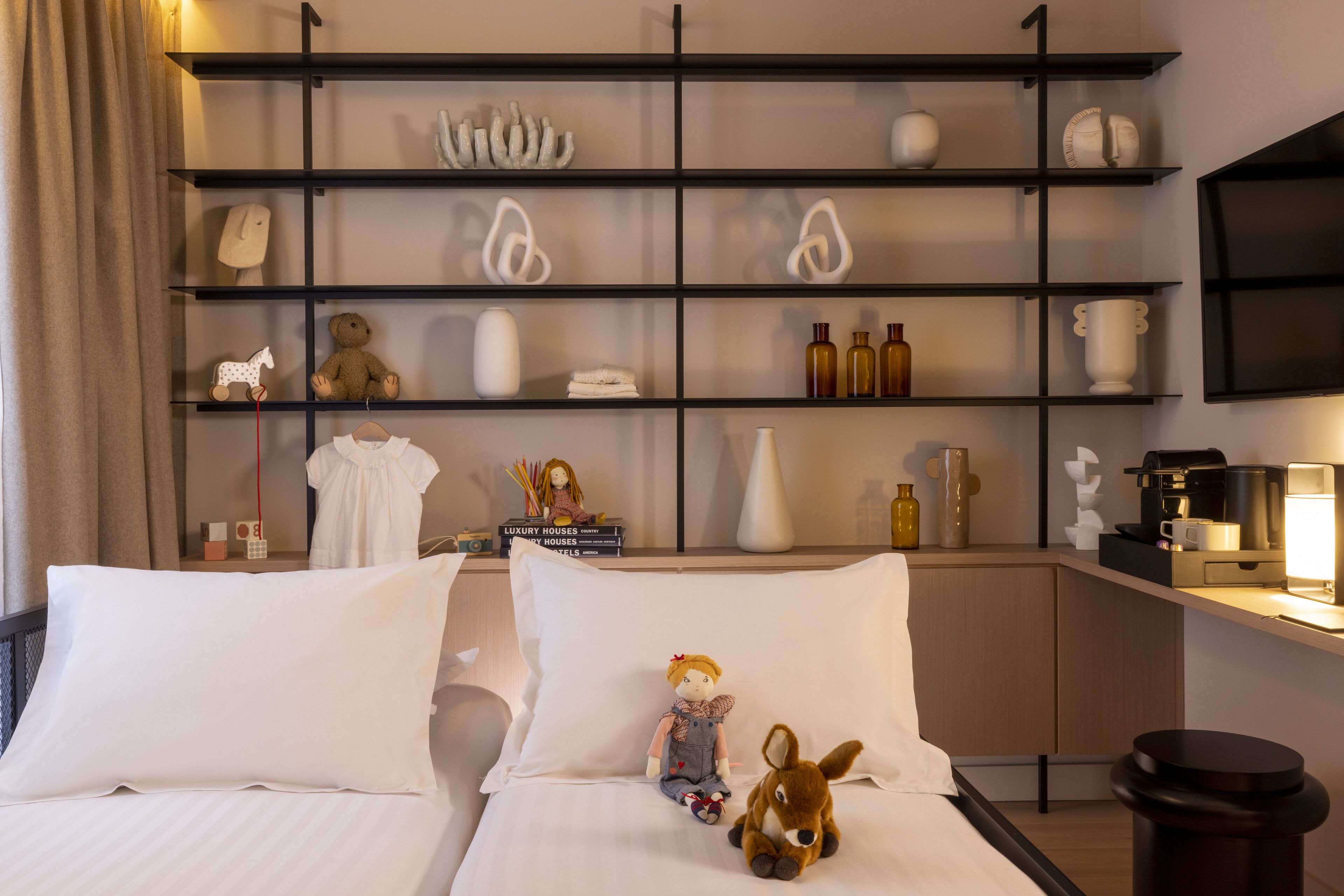 A hotel room with two beds, a shelf with decorative items, a coffee machine, and a TV on the wall.