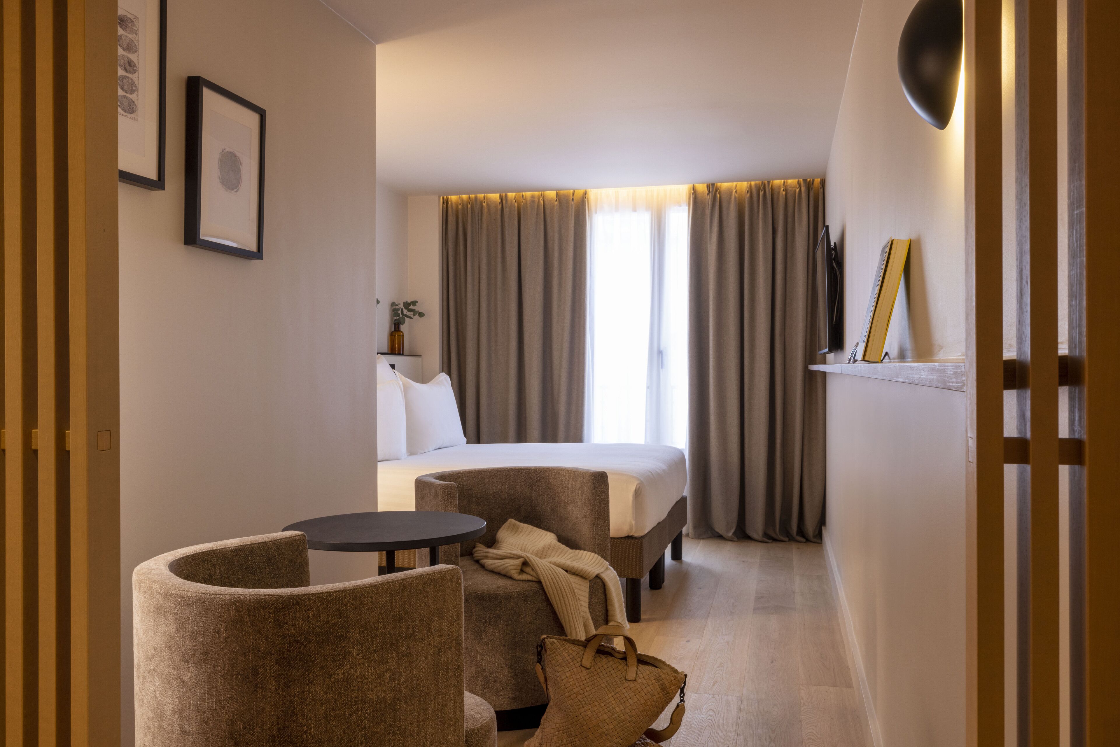 Een hotelkamer met een bed, twee fauteuils, een tafeltje, een nachtkastje met lamp en een raam met gordijnen.