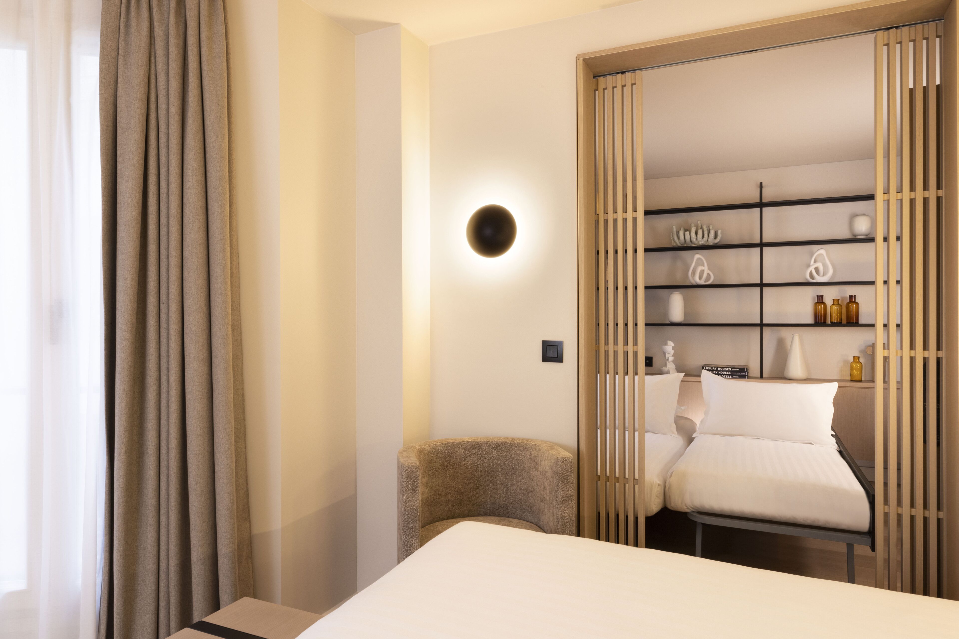 A modern hotel room with a bed, a chair, a wardrobe with shelves, and a wall-mounted light.
