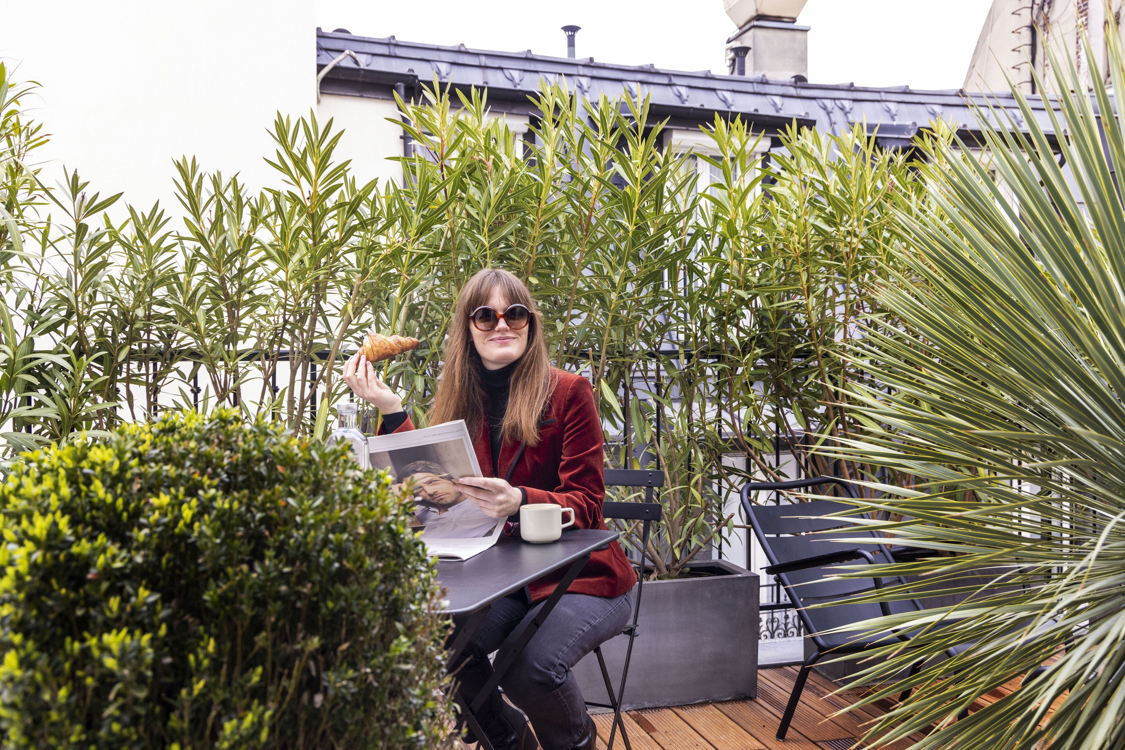 A person sitting at an outdoor table with a laptop and a cup, surrounded by greenery and a building in the background.