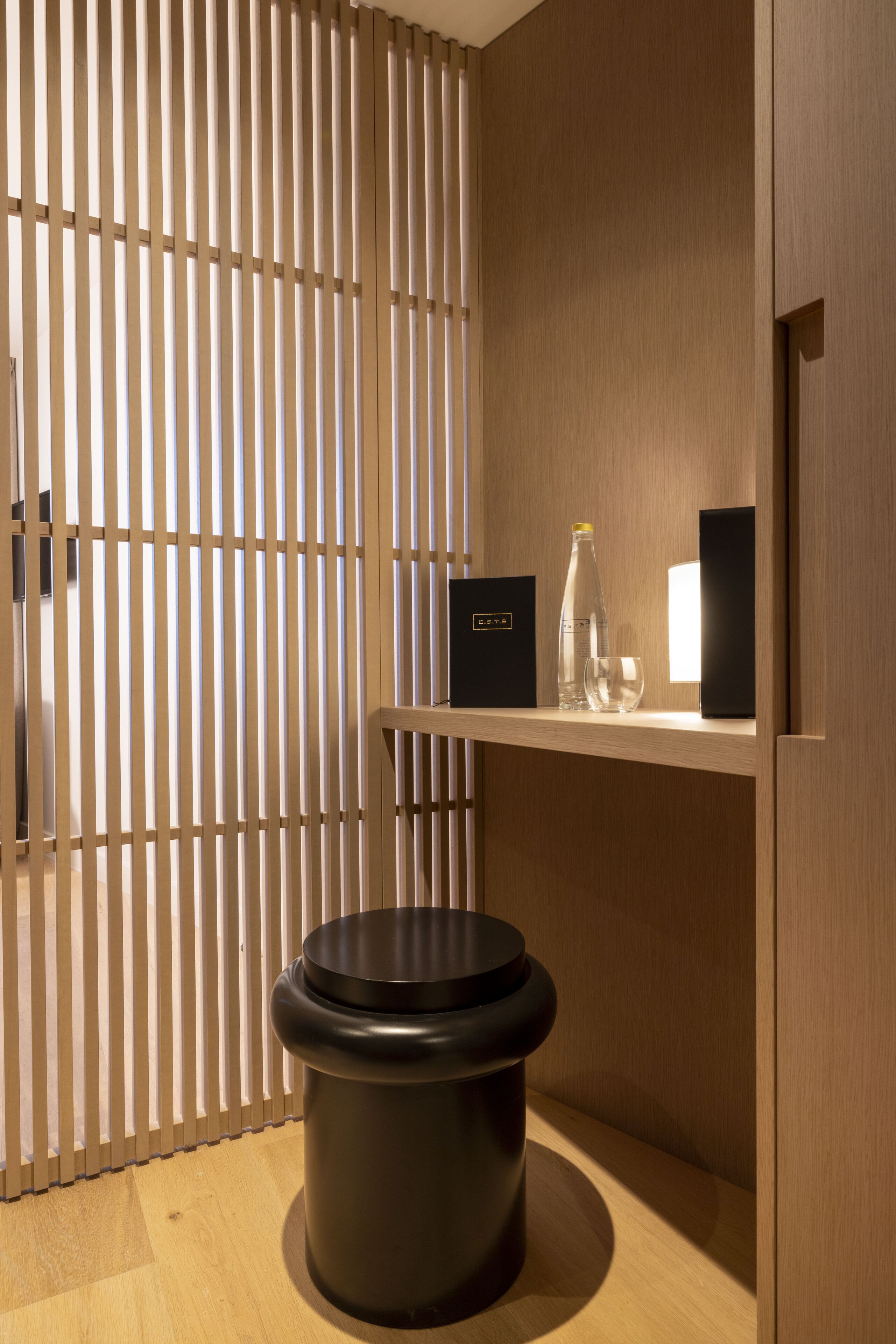 A modern bathroom with a wooden slatted partition, a black toilet, a shelf with a bottle, a glass, and a black box.