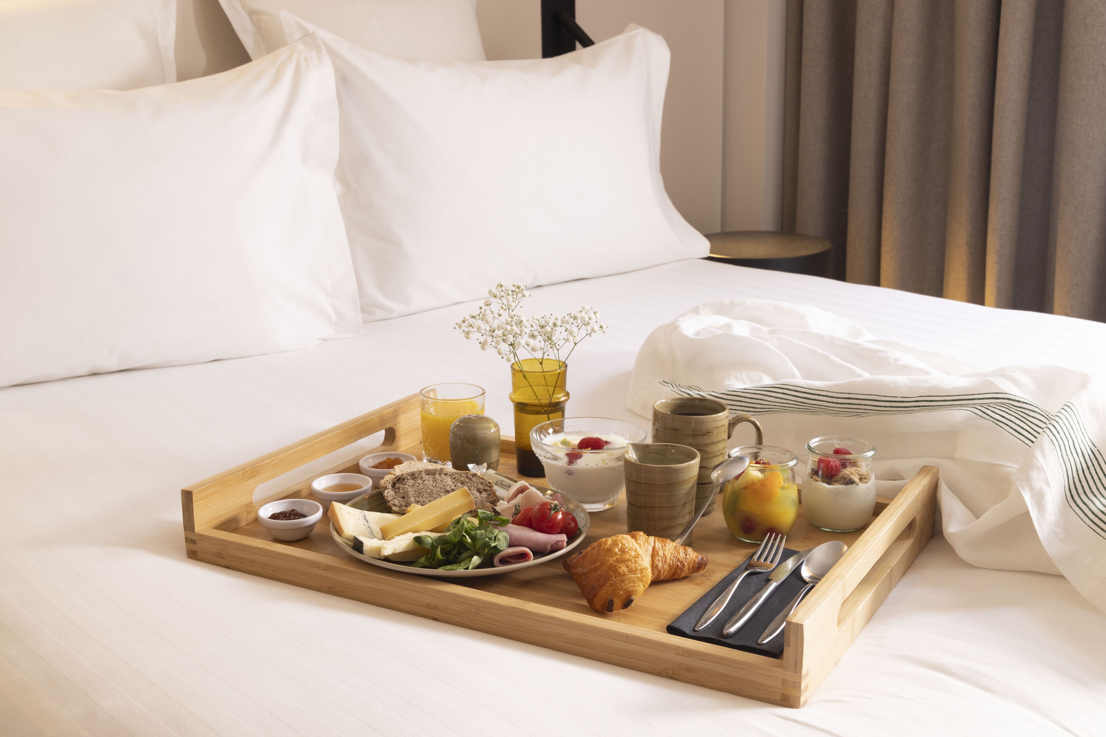A neatly arranged breakfast tray on a bed with white bedding, including a croissant, cheese, fruits, yogurt, and a small vase with flowers.