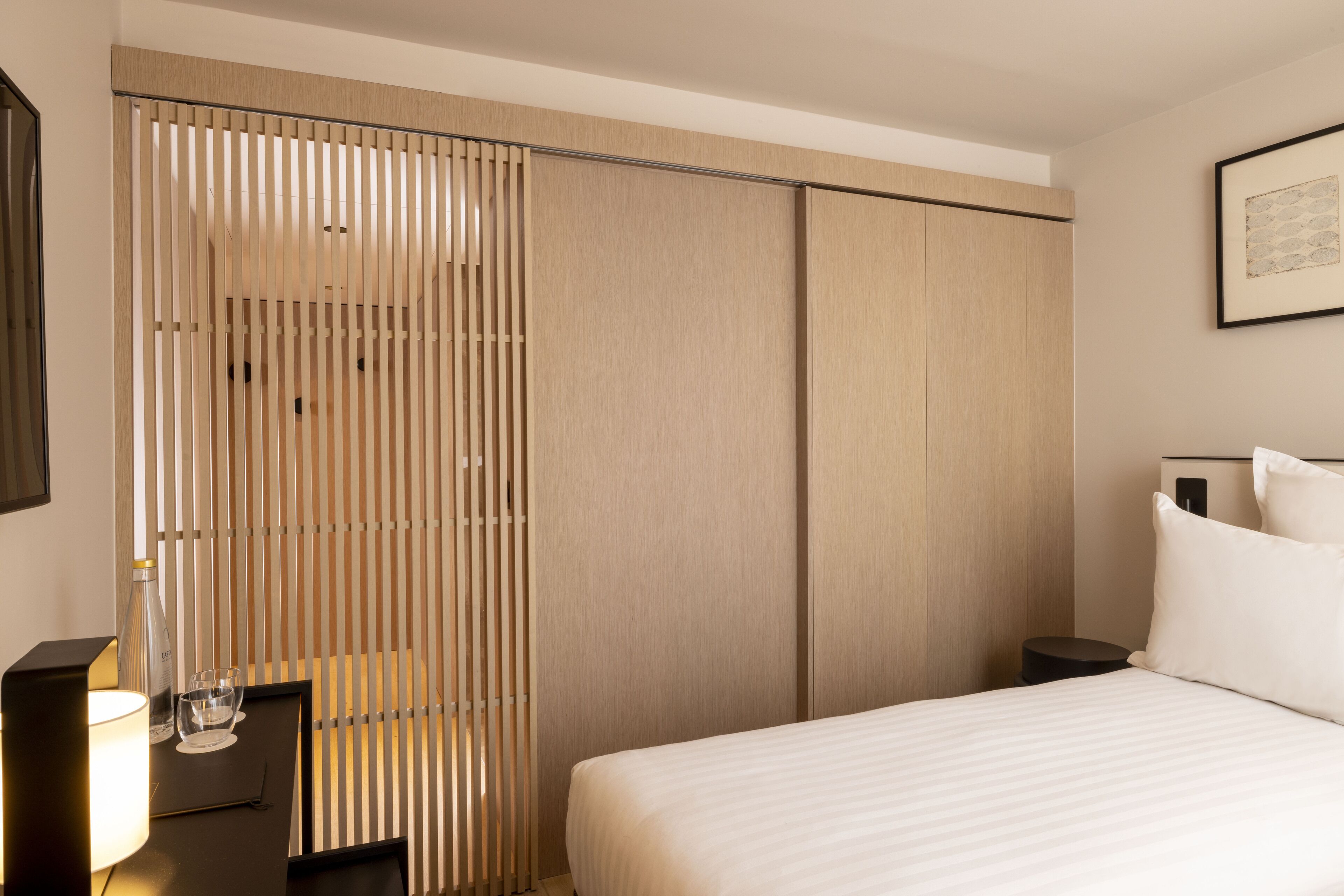 Une chambre d’hôtel comprenant un lit, des tables de chevet, une lampe et une porte coulissante avec des stores verticaux.