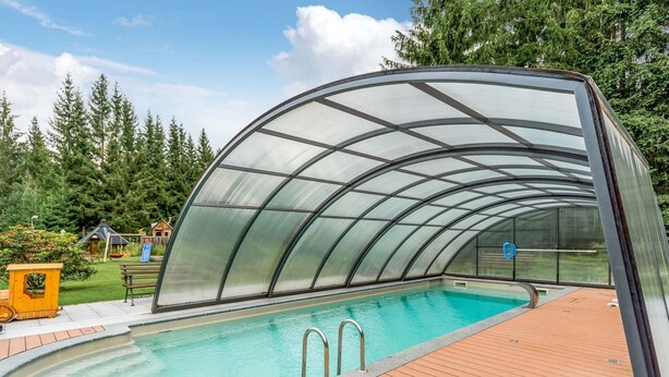 Ein Swimmingpool mit einem gläsernen Deck und einer umlaufenden Holzterrasse, umgeben von Grünpflanzen.