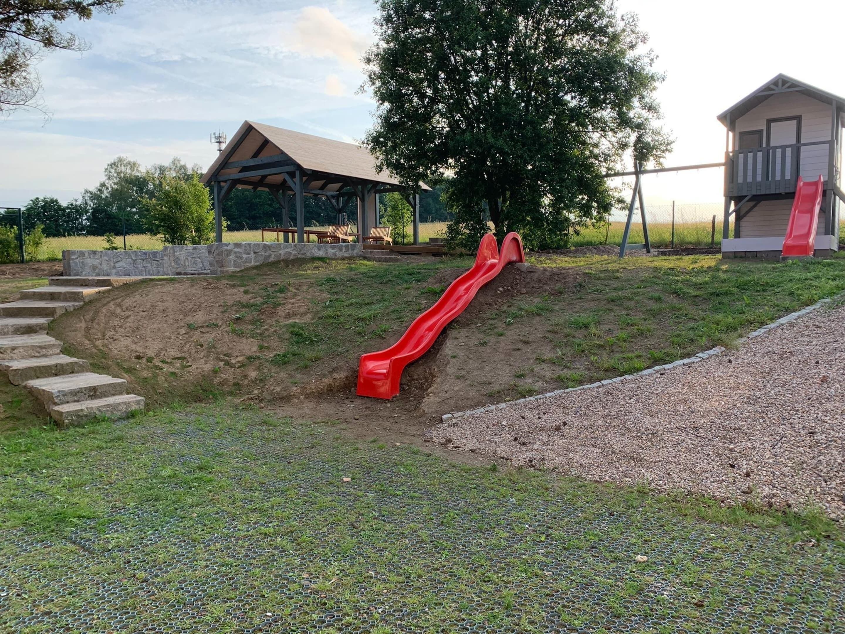 Une aire de jeux avec un toboggan rouge, un espace de repos abrité et un petit bâtiment également doté d’un toboggan rouge.
