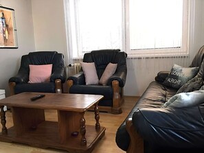 A living room with a wooden coffee table, two leather armchairs with cushions, a leather sofa, and a window with blinds.