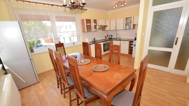 Uma área de refeições com mesa e cadeiras de madeira, cozinha ao fundo, e uma porta de vidro deslizante.