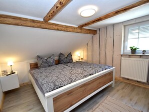 A bedroom with a sloped ceiling, exposed wooden beams, a bed with patterned bedding, bedside lamps, a window with blinds, and a radiator.