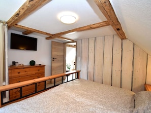 A bedroom with a sloped ceiling, wooden beams, a bed, a dresser, a TV, and a door leading to another room. - Ferienhaus mit Sauna & Spa, Huttenrode (Blankenburg)