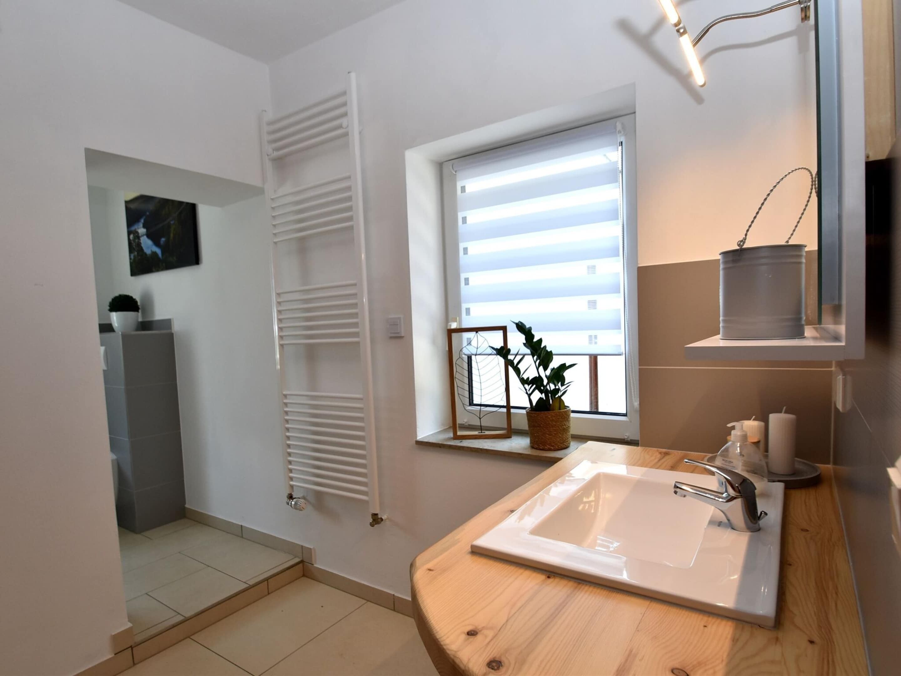 Une salle de bain avec un lavabo blanc, une fenêtre avec des stores, un porte-serviettes et une plante en pot.