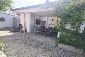 Relax outdoors with a dining table and TV, surrounded by lush greenery and an entrance.