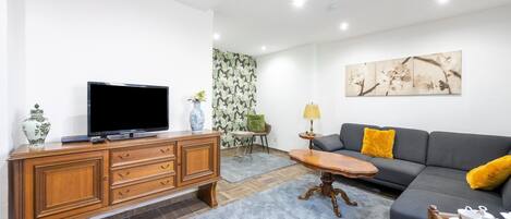 A living room with a flat-screen TV on a wooden cabinet, a grey sofa with yellow cushions, a small round table with a lamp, and a blue rug.
