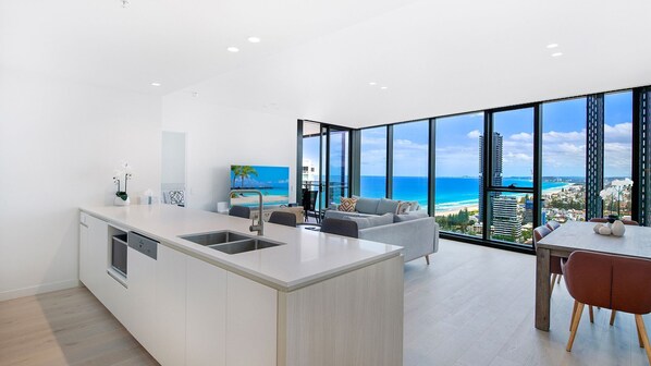 A modern kitchen with a large island, a dining area with a view of the ocean, and a living area with a sofa and a painting of a beach scene.