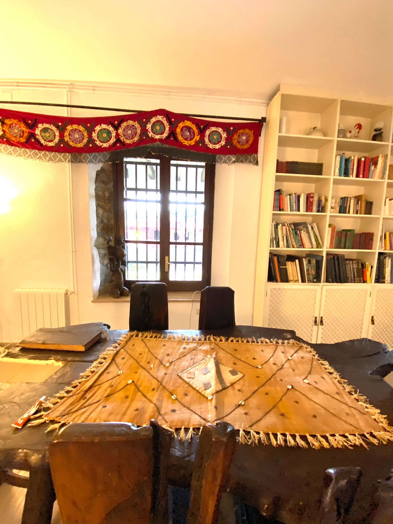 A room with a wooden table, two chairs, a bookshelf, and a window with decorative curtains.