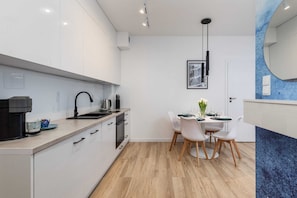 A modern kitchen with white cabinets, a black faucet, and a dining area with a white table and chairs. - Comfortable Apartment | Close to the Beach and Parking (Grzybowo)