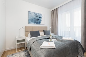A modern hotel room with a bed, a nightstand, a bedside table, a lamp, a painting, and a window with curtains. - Comfortable Apartment | Close to the Beach and Parking (Grzybowo)