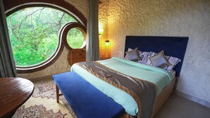 A bedroom with a large round window, a bed with a headboard, a bench, a table, and a patterned rug. - Chirping Hills Resort Bhimtal (Nainital)