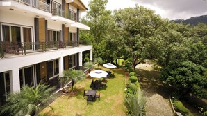 A modern hotel with a spacious outdoor area featuring a patio with tables and umbrellas, surrounded by lush greenery and trees. - Chirping Hills Resort Bhimtal (Nainital)