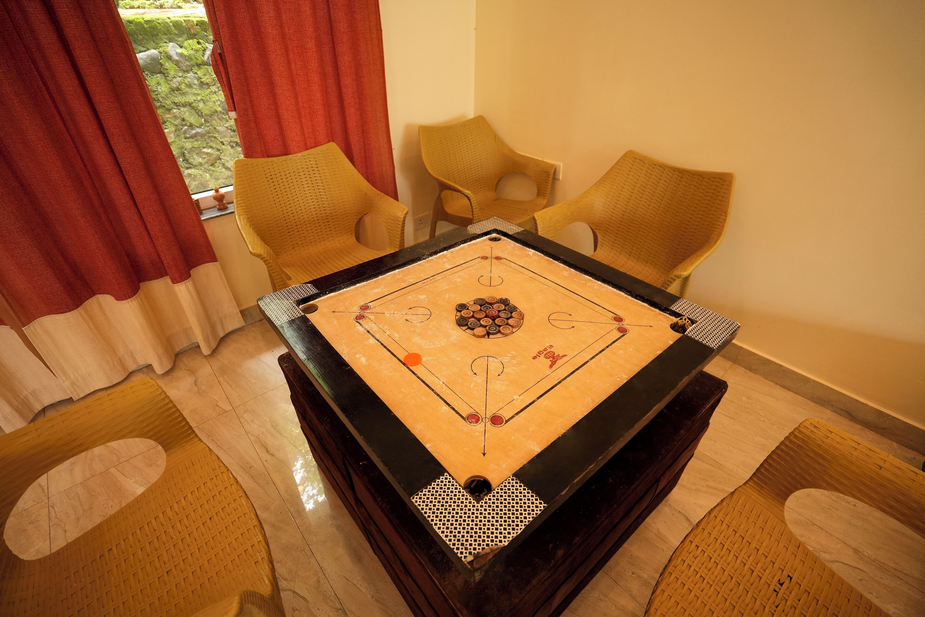 Una mesa de billar en una habitación con dos sillas de mimbre y un ventanal con cortinas rojas.