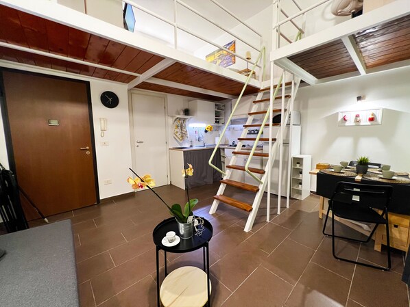 A spacious living area with a kitchen, a dining table with chairs, a staircase, and a small table with a plant and a cup.