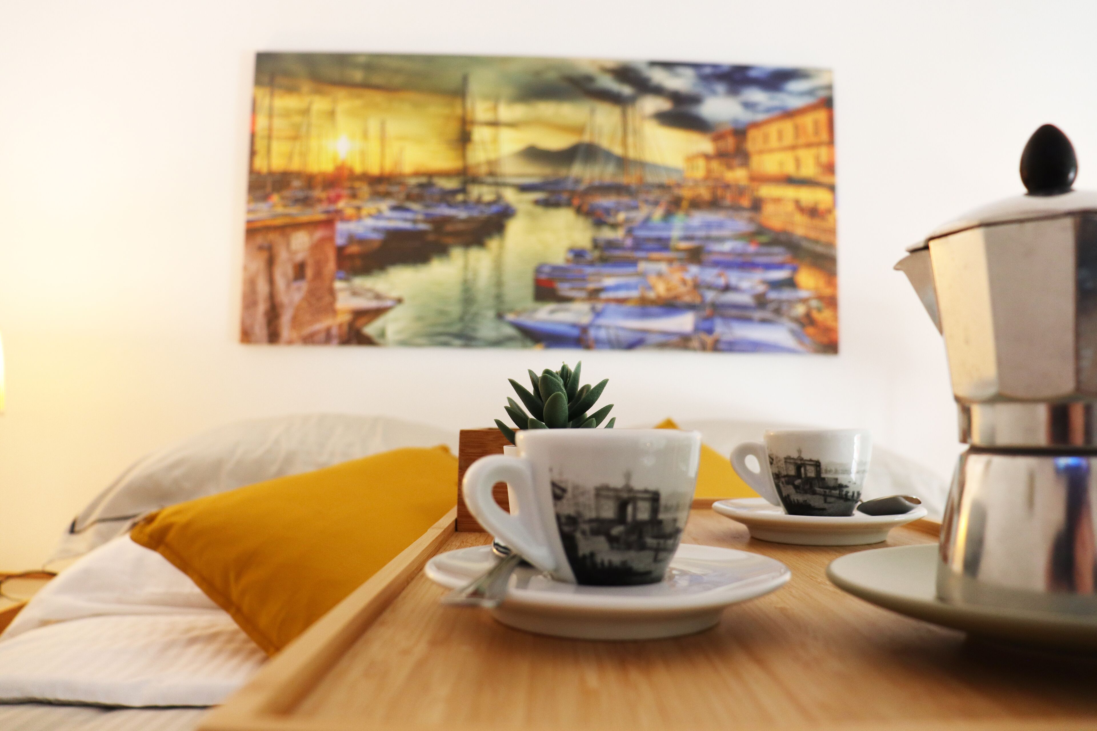 A neatly arranged breakfast table with a coffee pot, two cups, and a saucer, set against a wall with a painting of boats.