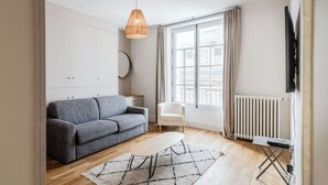 Ein modernes Wohnzimmer mit grauem Sofa, einem kleinen weißen Tisch, einer Hängelampe, einer Heizung und einem Fenster mit Vorhängen.