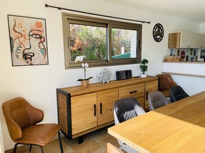 Ein Essbereich mit Holztisch und Stühlen, einer Holzkommode mit Dekorationselementen, einem Fenster mit Blick auf Bäume und einer Wanduhr.