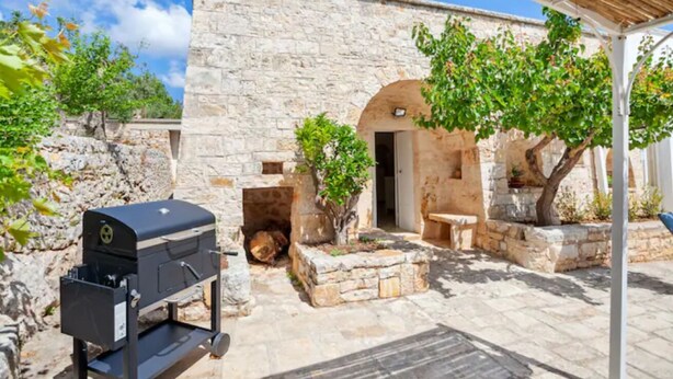 Un bâtiment en pierre surmonté d’un toit conique, doté d’un barbecue et d’un espace de repos couvert à l’ombre d’arbres.
