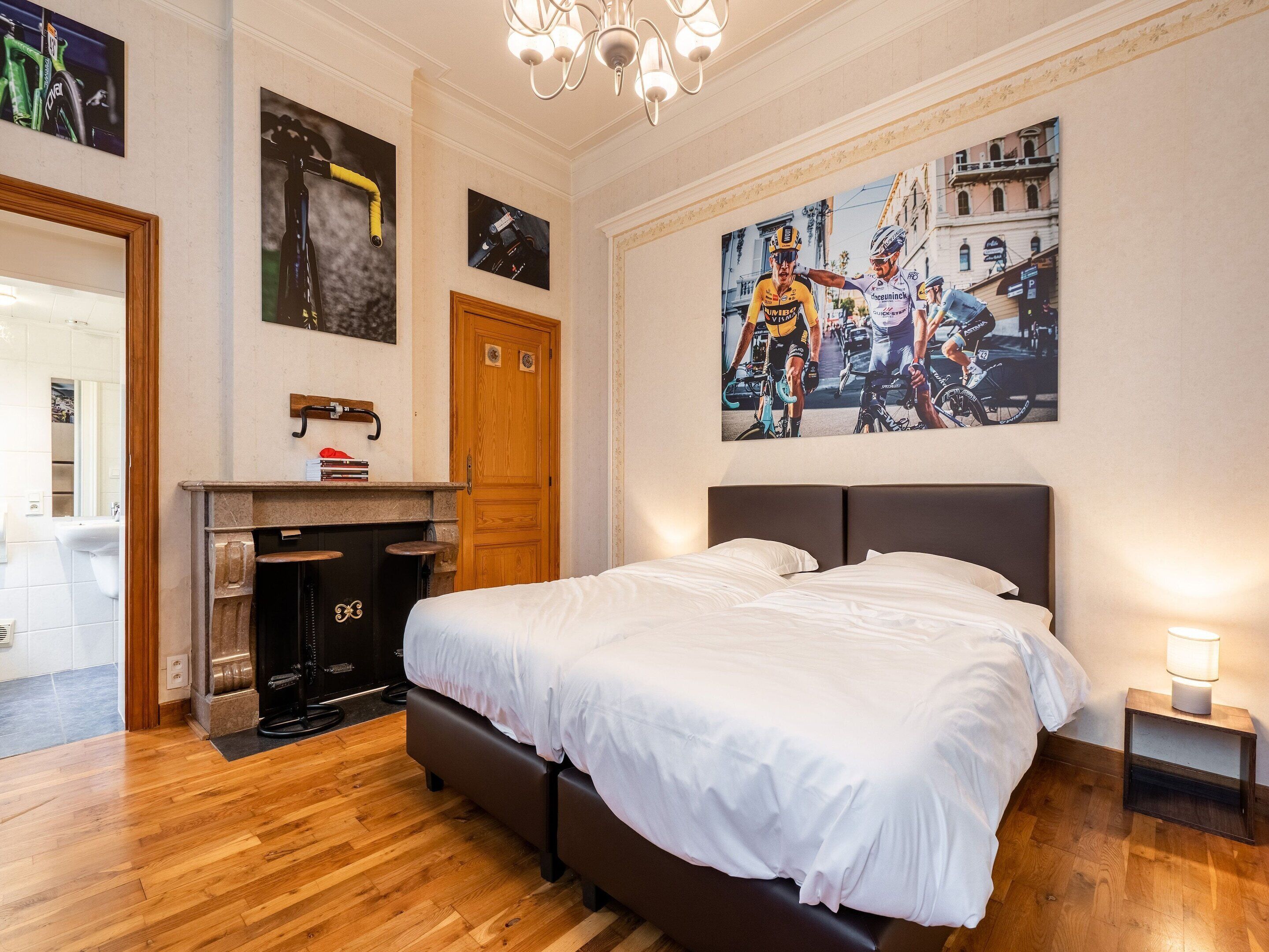 A bedroom with a large bed, a wooden floor, a fireplace, and a wall with cycling posters.