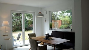 Ein Essbereich mit Holztisch, vier Stühlen, einer Couch, einer Pendelleuchte und Blick auf einen Garten durch große Fenster.