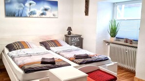 A bedroom with two beds, a nightstand, a lamp, a radiator, and a window with blinds. - Apartment in Deudesfeld With Private Terrace (Schutz)