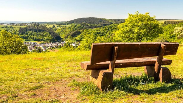 A wooden bench in a grassy field with a view of a village and hills in the background. - Apartment in Deudesfeld With Private Terrace (Schutz)