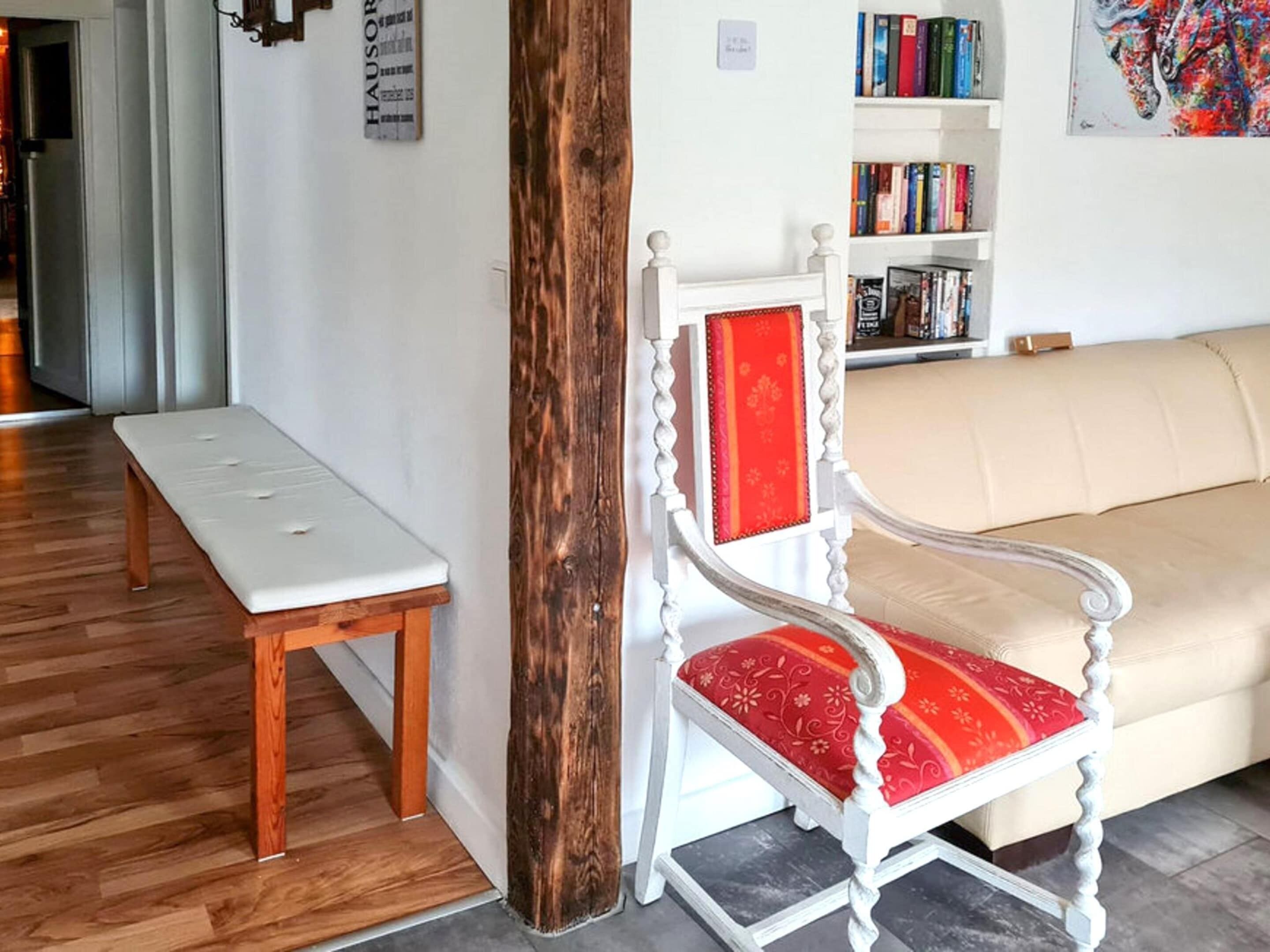 Ein Zimmer mit einer weißen Bank, einer Holzsäule, einem weißen Sessel mit rotem Kissen und einem Bücherregal voller Bücher.