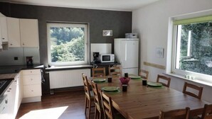 A modern kitchen with white cabinets, a microwave, and a refrigerator. There is a dining table with chairs and a view of greenery through the windows. - Holiday Home With Terrace in Sauerland (Brilon)