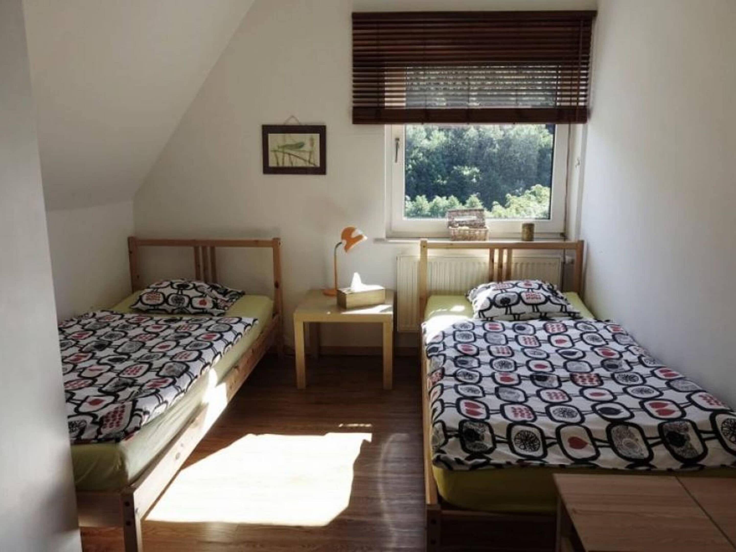 A small, well-lit room with two single beds, a small table, and a window with a view of greenery.