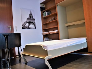 Ein kleines Zimmer mit einem Klappbett, einem Schreibtisch und einem Bücherregal.