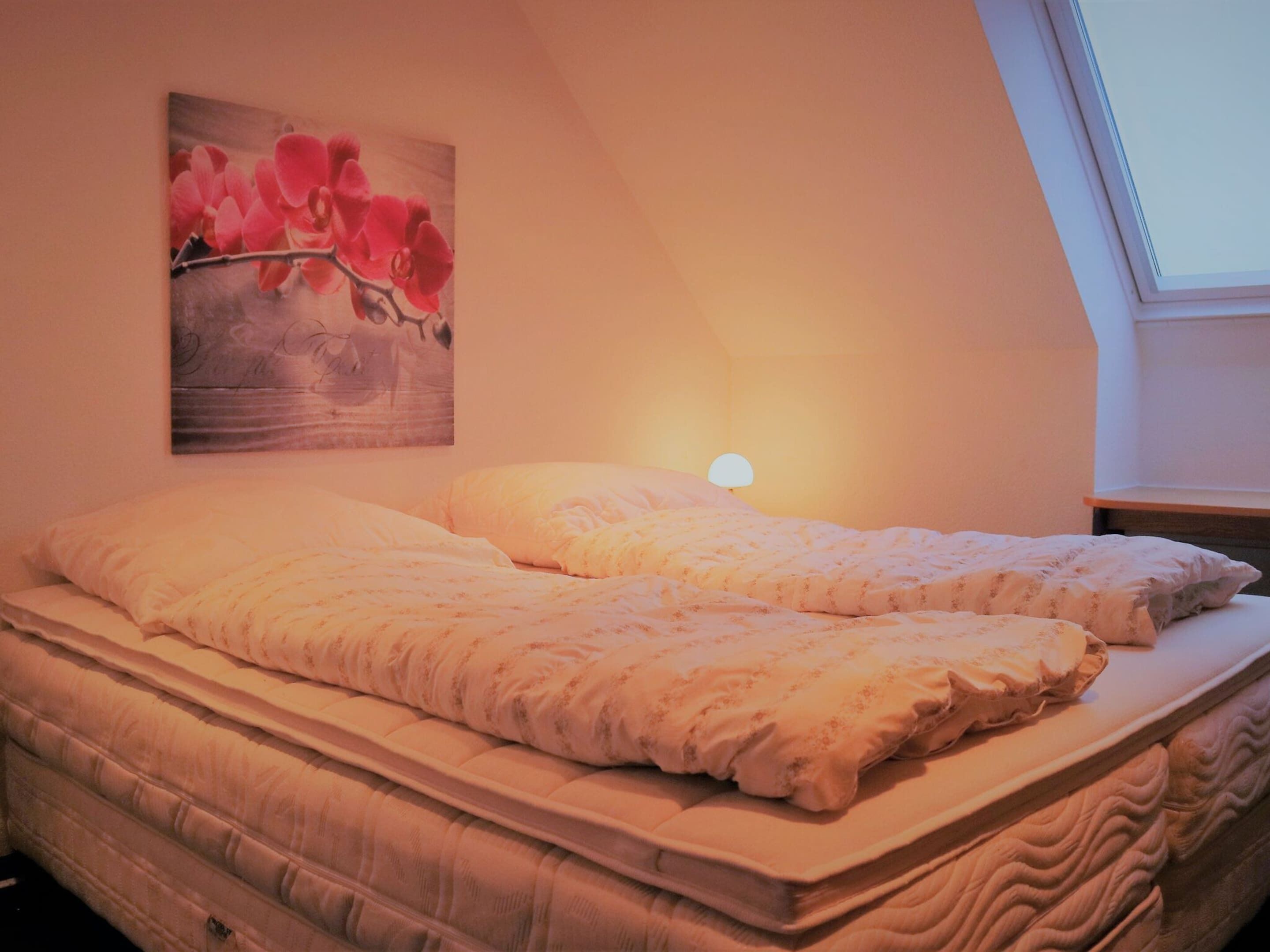 A bedroom with a bed, a nightstand, a lamp, and a painting of flowers on the wall.