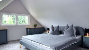 Un dormitorio con techo inclinado, una cama con ropa de cama gris, dos mesitas de noche y una ventana con vistas a árboles.