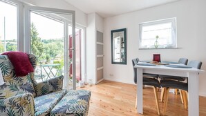 A modern living room with a sofa, dining table, and chairs. There is a large window with a view of greenery outside. - Pretty Holiday Home in Schleiden With Balcony and Barbecue (Schleiden)