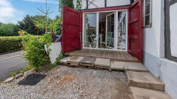 A traditional half-timbered building with a red door and glass sliding doors, a small outdoor seating area with a table and chairs, and a gravel pathway leading to the entrance. - Pretty Holiday Home in Schleiden With Balcony and Barbecue (Schleiden)
