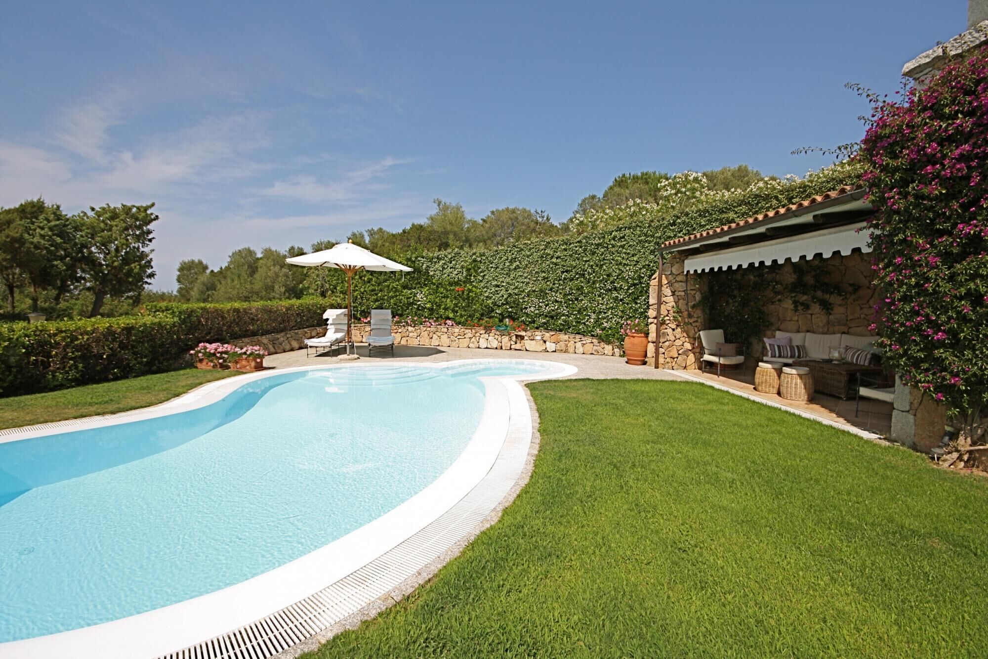 Una piscina con un muro in pietra e un prato. Sono presenti lettini e un tavolo con sedute all'aperto.