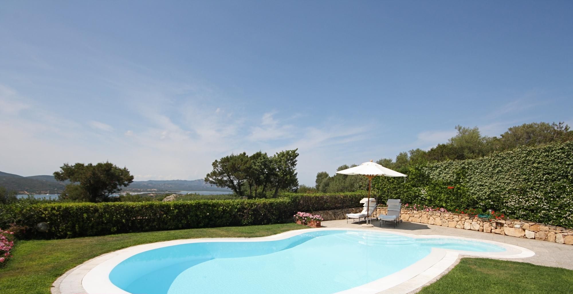A swimming pool with a white deck, surrounded by a well-maintained lawn and a stone wall.