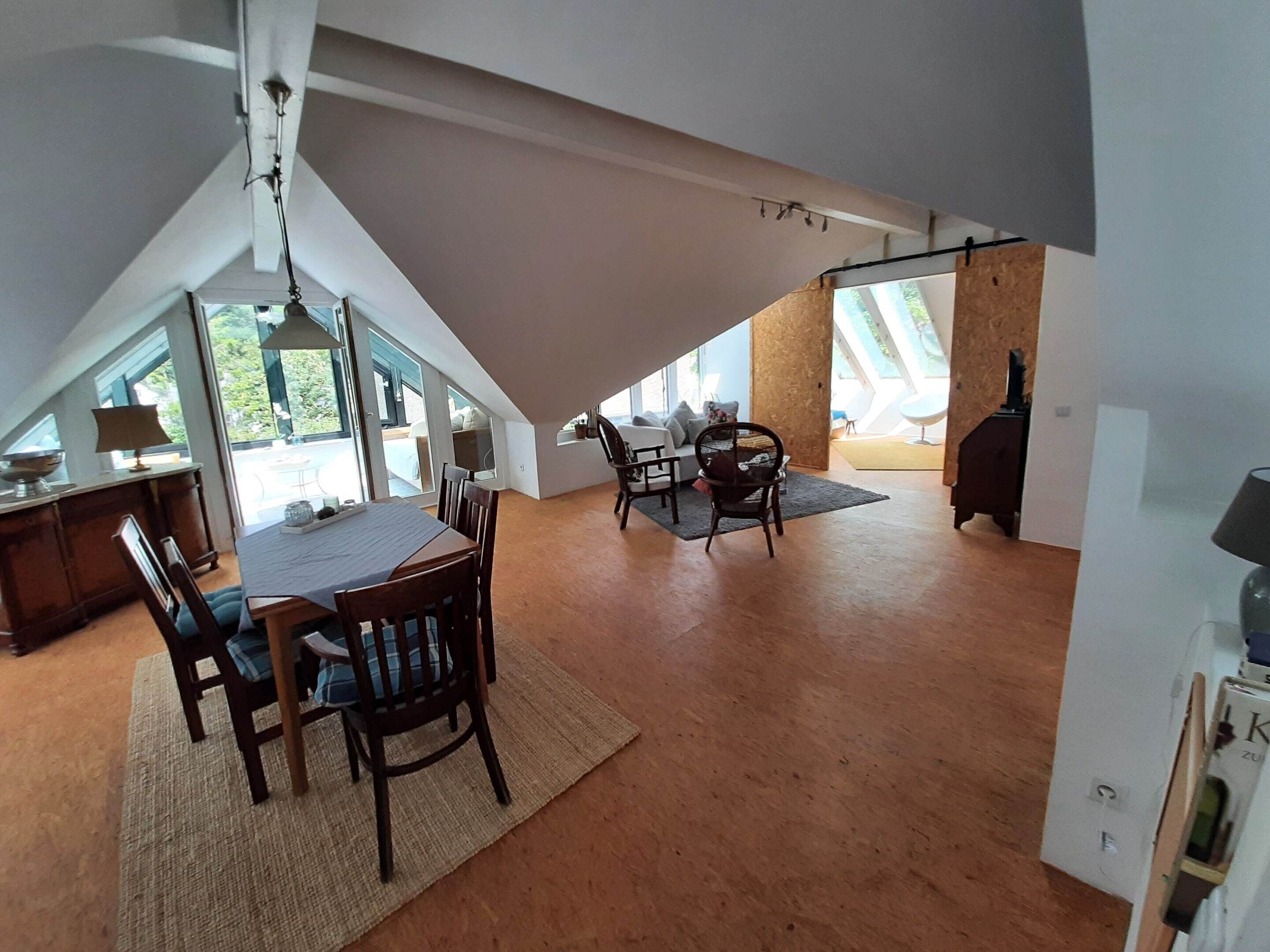 A spacious loft with a high ceiling, large windows, and a dining area with wooden furniture.