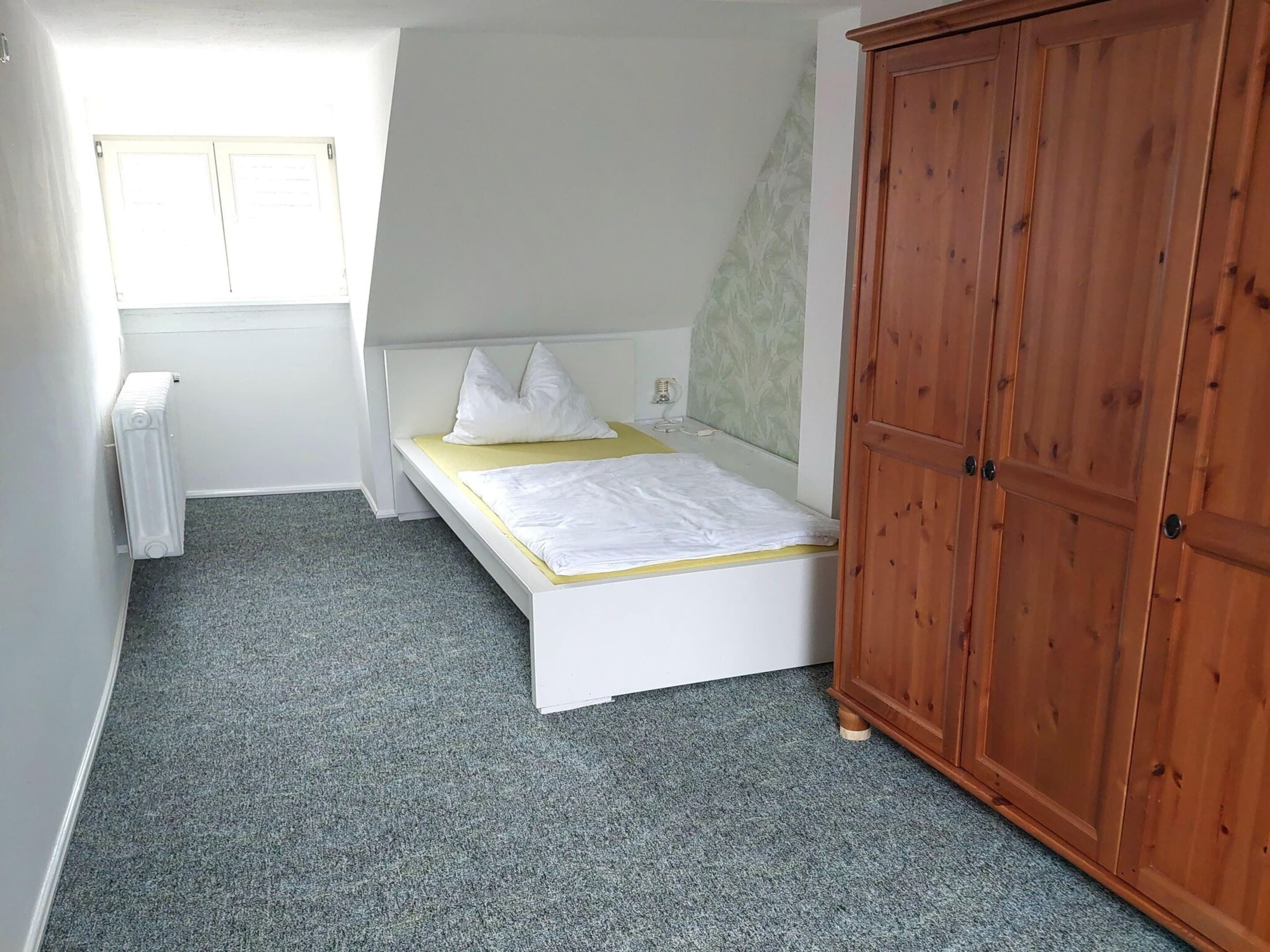 Une petite chambre avec un lit simple, une armoire en bois, un radiateur et une fenêtre.