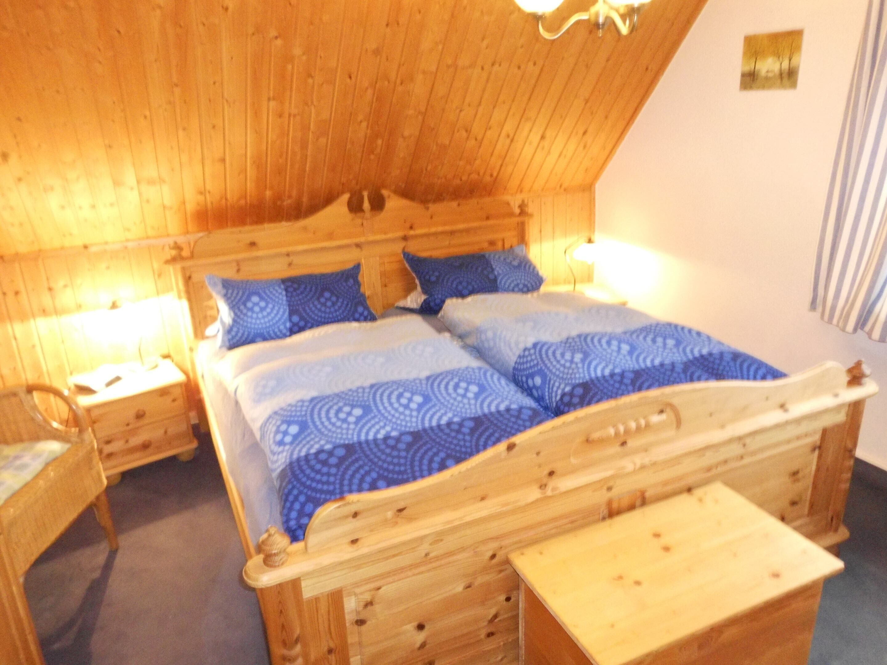 A wooden four-poster bed with blue patterned bedding, two bedside tables, and a small wooden bench.