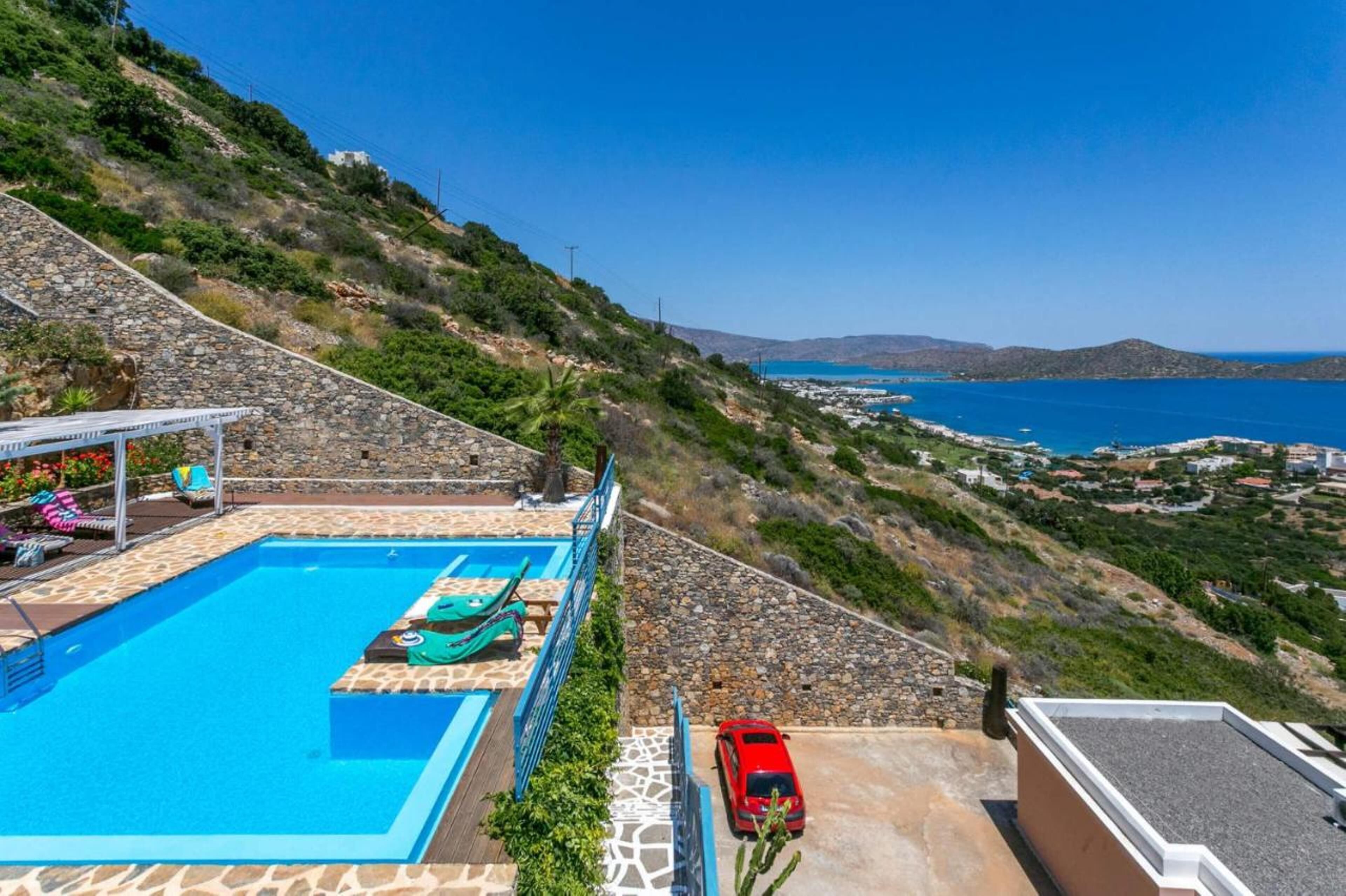 Uma piscina com um deck de pedra, um carro vermelho e vista para o mar e colinas.
